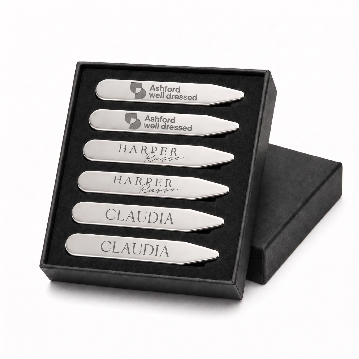 Six custom metal collar stays with engraved logos arranged in a black presentation box